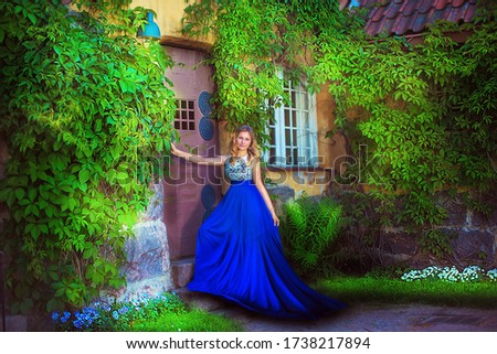 blonde girl in a blue dress standing in the park at the door of the house