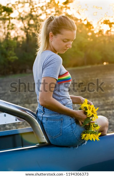 The blonde girl in\
denim skirt sits on the trunk of the car with a bouquet of\
sunflowers in her hands