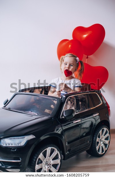 Blonde girl eating a caramel Lollypop in the\
shape of a heart. Kid sitting in a black toy car with red heart\
shaped baloons.Valentine\'s Day\
concept