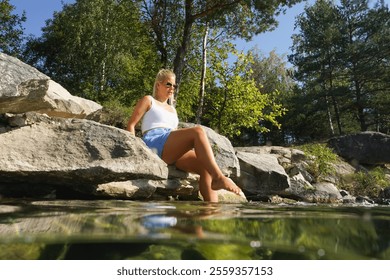 A blonde girl of European appearance wets her feet while sitting on the rocky shore of a lake on a summer day. - Powered by Shutterstock