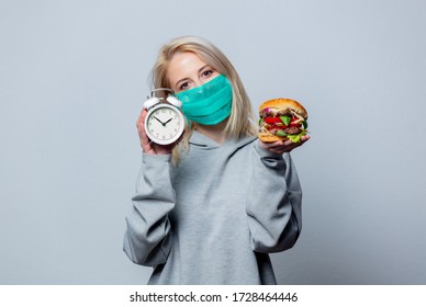 Blonde Girl In Face Mask With Burger And Alarm Clock On White Background