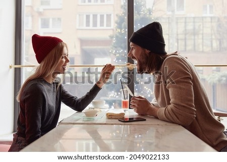 Blonde girl feeding her bearded boyfriend in modern cafe. Concept of romantic relationship. Comfortable lifestyle. Young european couple wearing hats and casual clothes. Persons sitting at table
