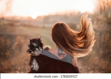 A blonde girl with fluttering hair stands on her back with the tattoo of her black Schnauzer dog, which she holds in her arm. Pet portrait. Autumn Travel, walking with pets. Close up portrait, mock up