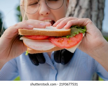 Blonde girl with glasses is resting to eat fast food in the park. Close-up of a beautiful female student having lunch with a sandwich outdoors.