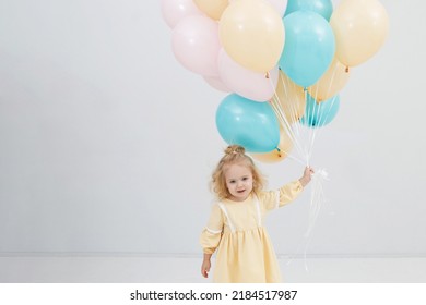 Blonde girl has fun and rejoices with a bunch of green and yellow balloons in a white studio. Festive decoration. Children's happiness.