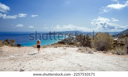 Blonde girl with hat walking on rocky ledge facing the coastline of Zante, Greece in a summer day