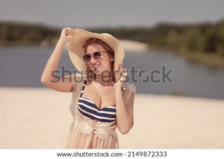 A blonde girl with long hair in a white hat is resting on the white sand near the sea on a sunny day