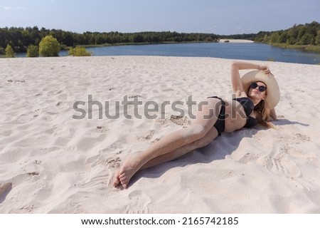 A blonde girl with long hair in a white hat is resting on the white sand near the sea on a sunny day