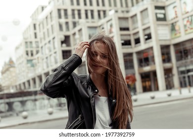 Blonde Girl With Long Windy Hair. Portrait Of The Girl In Black Faux Leather Jacket On The Building Background