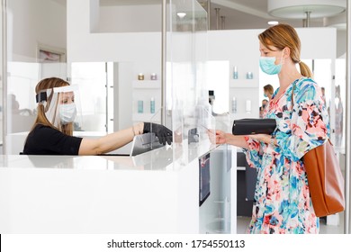 Blonde Girl With Mask Pays At The Checkout With A Credit Card Where The Cashier Is Protected By A Plexiglass Plate And A Visor With Mask