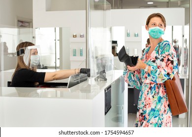 Blonde Girl With Mask Pays At The Checkout With A Credit Card Where The Cashier Is Protected By A Plexiglass Plate And A Visor With Mask