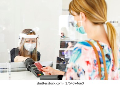 Blonde Girl With Mask Pays At The Checkout With A Credit Card Where The Cashier Is Protected By A Plexiglass Plate And A Visor With Mask