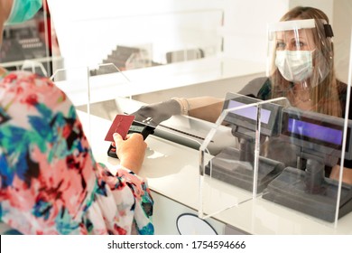 Blonde Girl With Mask Pays At The Checkout With A Credit Card Where The Cashier Is Protected By A Plexiglass Plate And A Visor With Mask