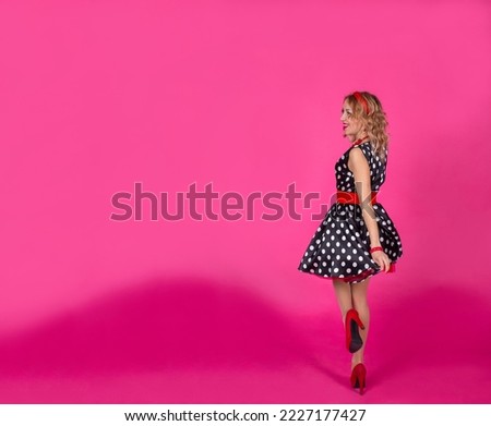 A blonde girl on a pink background in a black dress with white polka dots with red accessories: high heel shoes, beads with a bracelet and earrings, a hoop on her head. Lips with red lipstick. Pin-up.