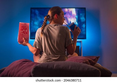 A blonde girl in pajamas watches TV at night. Insomnia. She is holding a pack of popcorn and a carbonated drink with a straw. Neon light. Watching TV shows and series, quarantine.