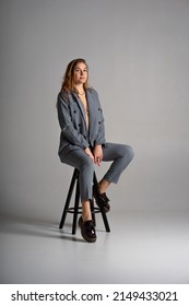 A blonde girl in a pantsuit with an unbuttoned jacket from under which can see a beautiful slender sexy body and underwear and black platform shoes poses in the studio sitting on a bar stool on white