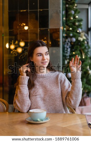 blonde girl in a pink sweater in a cafe on a background of Christmas trees and Christmas lights bokeh 
waving hello with a cup of coffee