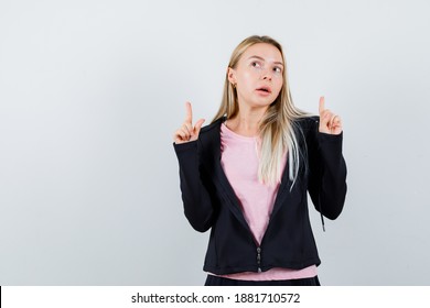  blonde girl pointing up in pink t-shirt and black jacket and looking cute. front view.  - Shutterstock ID 1881710572