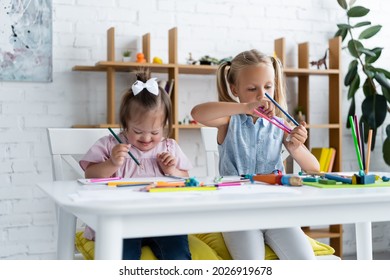 Blonde Girl Reaching Color Pencils Near Disabled Toddler Child With Down Syndrome In Private Kindergarten 