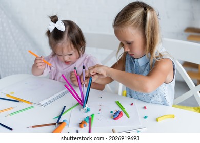 Blonde Girl Reaching Color Pencils Near Disabled Toddler Kid With Down Syndrome In Private Kindergarten 