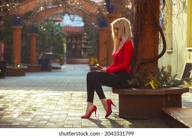 A blonde girl in a red blouse and red heels looks at the mobile phone