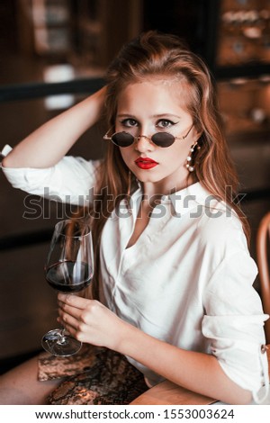 blonde girl with red lips in a white blouse in a snake skirt in sunglasses with a glass of wine in a restaurant in a relaxing atmosphere