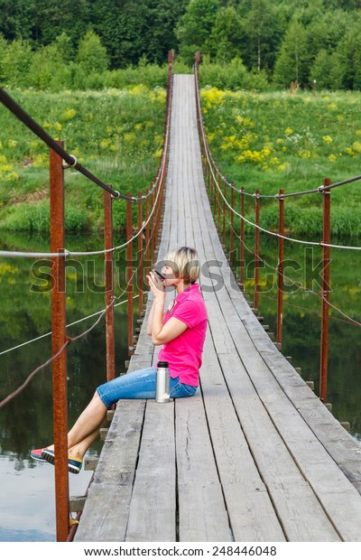 blonde girl with short haircut drinking tea from a\
thermos on Pedestrian suspension bridge of steel and wood over the\
river, summer in Russia