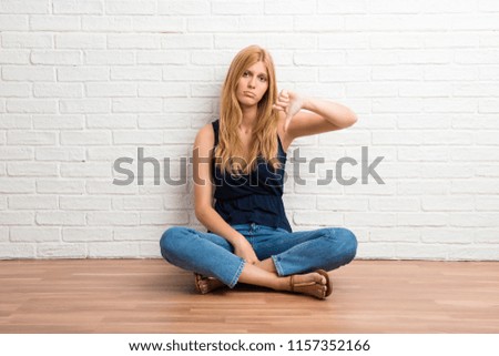 Blonde girl sitting on the floor showing thumb down sign with negative expression on white brick wall background