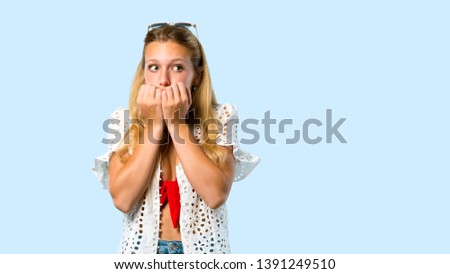 Blonde girl in summer vacation is a little bit nervous and scared putting hands to mouth on blue background