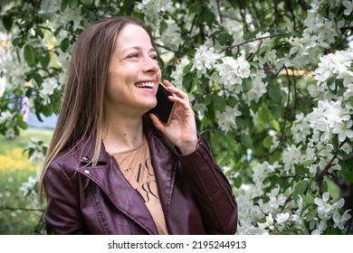 A Blonde Girl Walking In The Park Speaks On A Smartphone And Laughs, The Spring Season Is The Blossoming Of The Apple Tree, People Use Technology.