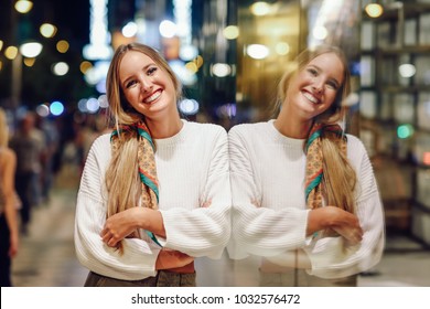 Blonde girl wearing white sweater smiling in the street with defocused city lights at the background. Pretty woman with pigtail hairstyle at night reflected in a shop window. - Powered by Shutterstock