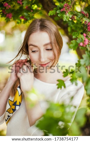a blonde girl in a white sweatshirt olive trousers with a brown leather bag with a yellow handkerchief in her hair in nature in the greenery in spring in a blooming garden trees posing walking