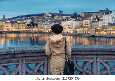 Blonde girl with wool hat and heavy clothing staring at the Limmat river in central Zurich at dusk in winter