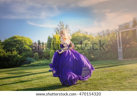 Blonde gorgeous woman in long purple luxurious dress posing in garden. Dress fluttering on wind.