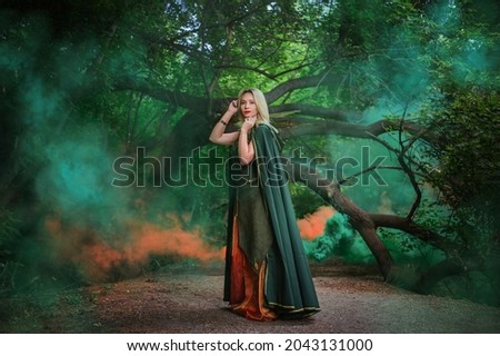 A blonde in a green cloak of a druid and in a fantastic dress with leaves in the fog. A sorceress girl in medieval dress stands in the forest in green and orange smoke. The image of a good witch