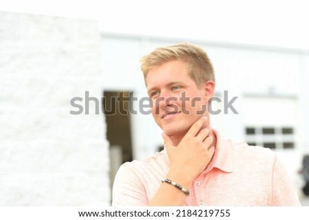 Blonde Hair Blue Eyes Caucasian American Man Smiling with a Hand and Face Pose in a Light Coral Shirt and Beaded Bracelet Touching his Attractive Jawline
