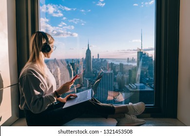 Blonde hair female blogger listening to music in headphones during freelance work on laptop computer. Hipster girl traveler enjoying panoramic view of New York downtown and studying with audio book - Shutterstock ID 1321453508