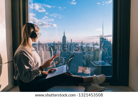 Blonde hair female blogger listening to music in headphones during freelance work on laptop computer. Hipster girl traveler enjoying panoramic view of New York downtown and studying with audio book