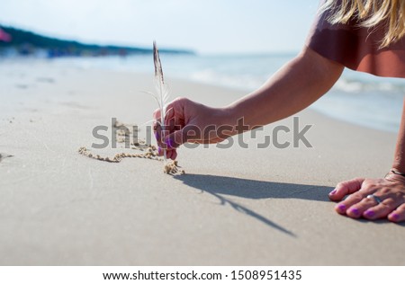 Blonde hair young woman is writting with a bird feather on sand. Girl drawing a heart on the sand at sunset in a summer love concept. A girl writing with sands with the sea background