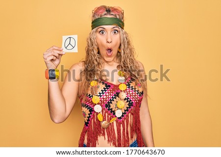 Blonde hippie woman wearing sunglasses and accessories holding reminder with peace symbol scared and amazed with open mouth for surprise, disbelief face