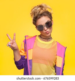 Blonde Hipster Girl With Hair Bun Wearing Colorful Jacket, Yellow Sheer Shirt Grimacing Making V-sign. 