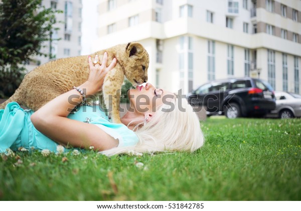 Blonde kisses calf of lion and lies on grass near\
building at summer day