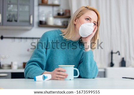 Blonde lady in mask with cup of hot tea and thermometer at kitchen table 