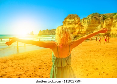Blonde lifestyle female tourist with raised arms on Praia Dona Ana at sunset. Caucasian woman enjoying at popular Dona Ana Beach in Lagos, Algarve coast. Summer holidays in Europe. Tourism in Portugal