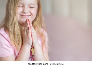 A blonde little girl, smiling, praying at home in the morning with folded hands and holding a cross. Children's prayer, faith and prayer to God.