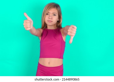 Blonde Little Kid Girl Wearing Pink Sport Clothes Over Green Background Feeling Unsure Making Good Bad Sign. Displeased And Unimpressed.