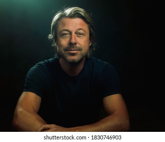 Blonde Man With A Grin Wearing Black T-shirt In Front Of Dark Background Looking At Camera.