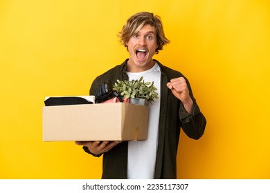blonde man making a move while picking up a box full of things isolated on yellow background celebrating a victory in winner position