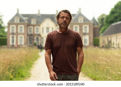 Blonde Man With A Stubble Beard In A Brown T-shirt On A Sandy Path Near A Country House In Summer. Front View.