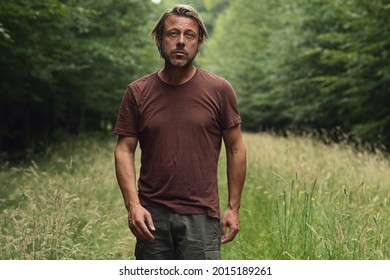 Blonde Man With A Stubble Beard In A Brown T-shirt Walks On A Forest Path Overgrown With Tall Grass In The Summer. Front View.